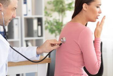 Breathing System Check Up Concept. Male general practitioner examining coughing female patient in modern clinic, using stethoscope, listening lungs from the back. Medicine And Healthcare